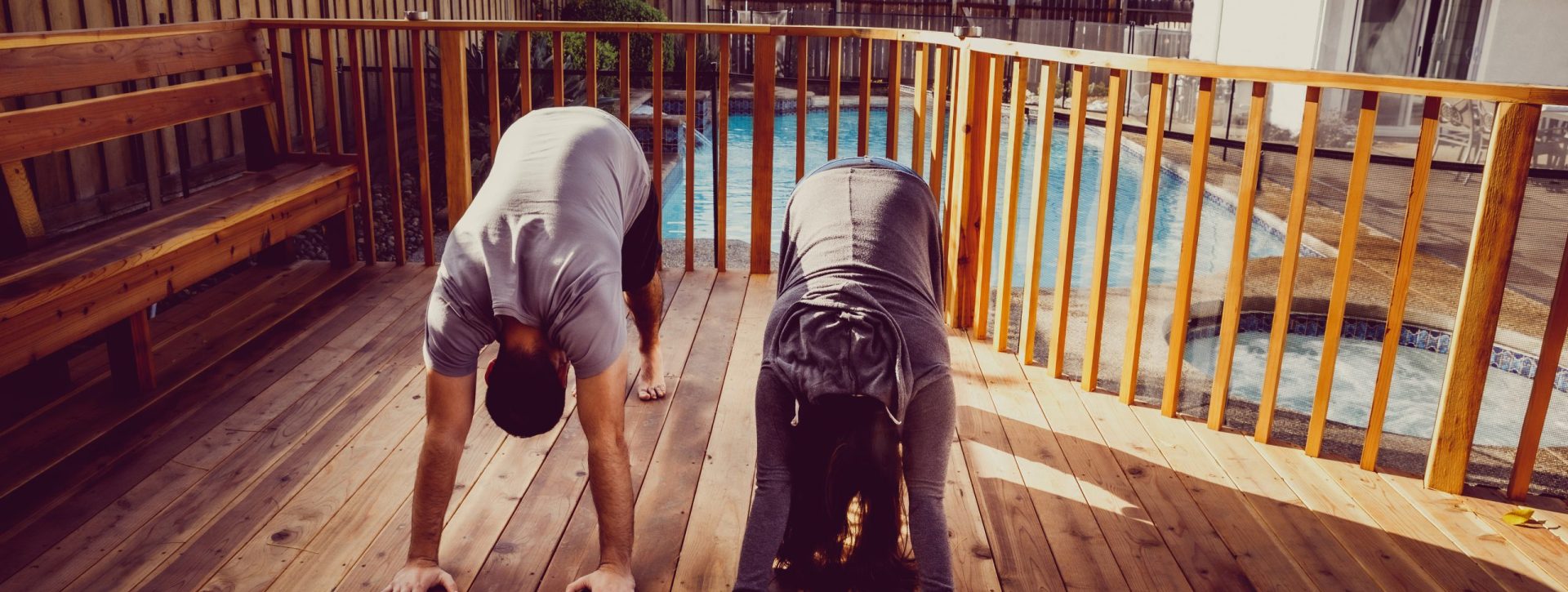 Two people in a downward dog pose while practicing yoga on a deck in front of a pool